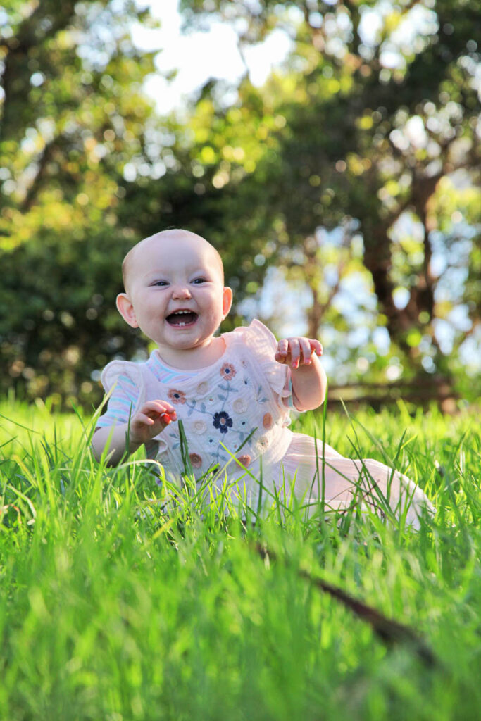 Smiling baby sitting in tall green grass during a sunny outdoor family photoshoot with Southern Exposure Photography in the Sutherland Shire