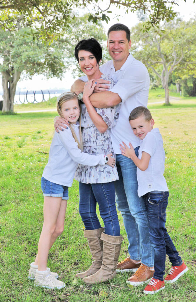 Family of four embracing outdoors under the trees, with parents and two young children smiling during a natural photoshoot with Southern Exposure Photography in the Sutherland Shire