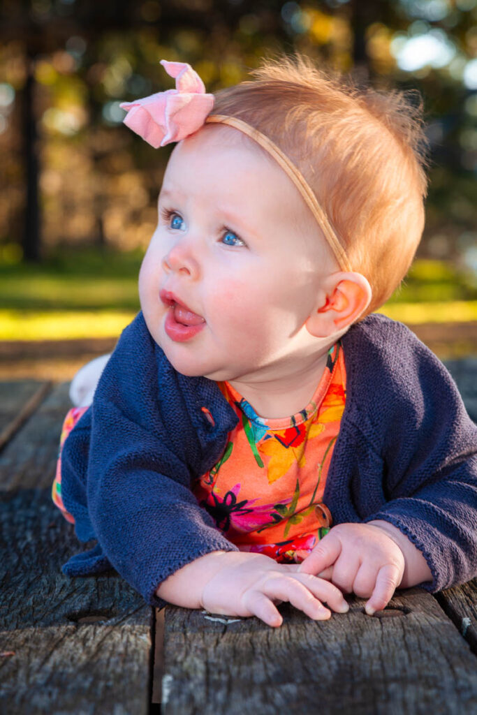 Baby with a pink bow headband and bright floral outfit lying on a wooden bench outdoors during a family photoshoot with Southern Exposure Photography